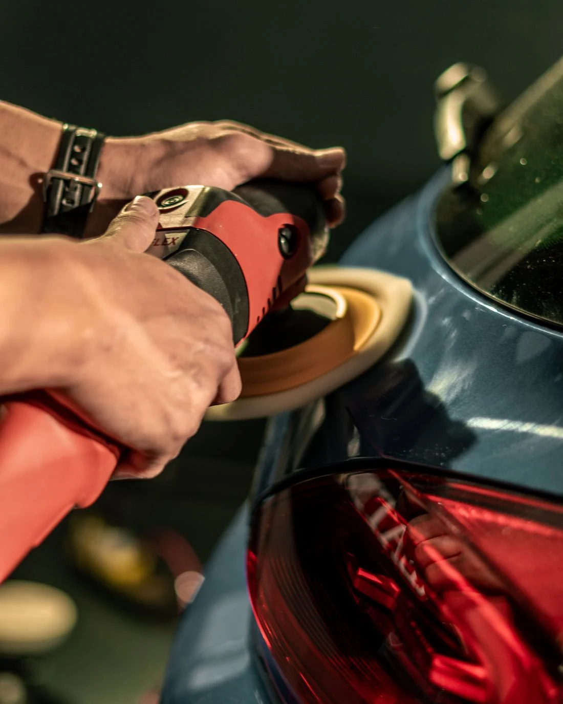 Hands guiding a polishing machine over a car's rear panel beside the tail light