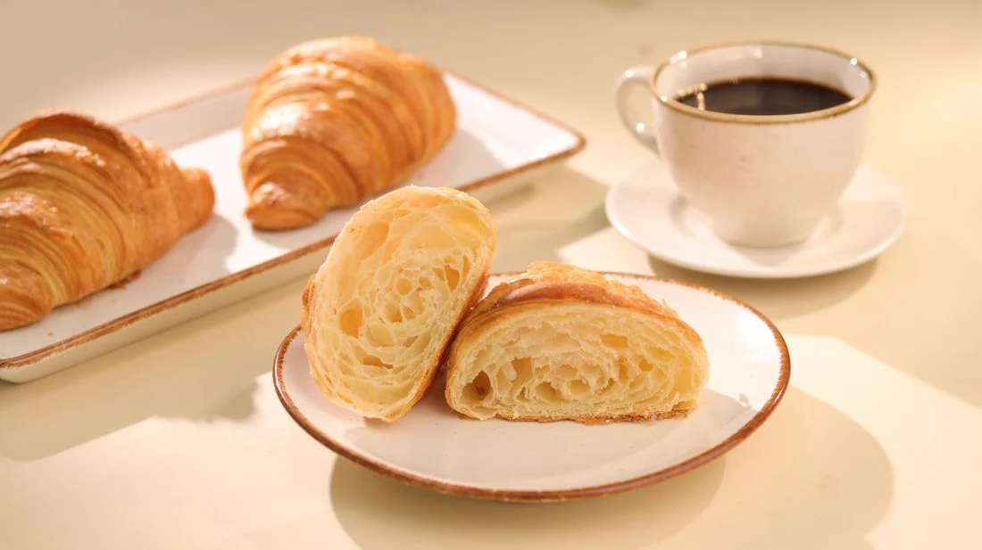 Golden croissants, one cut open to show the flaky layers, served beside a cup of coffee